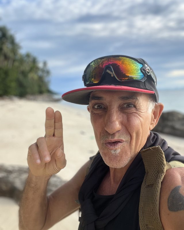Robin Good on a beach, cap on, two fingers up